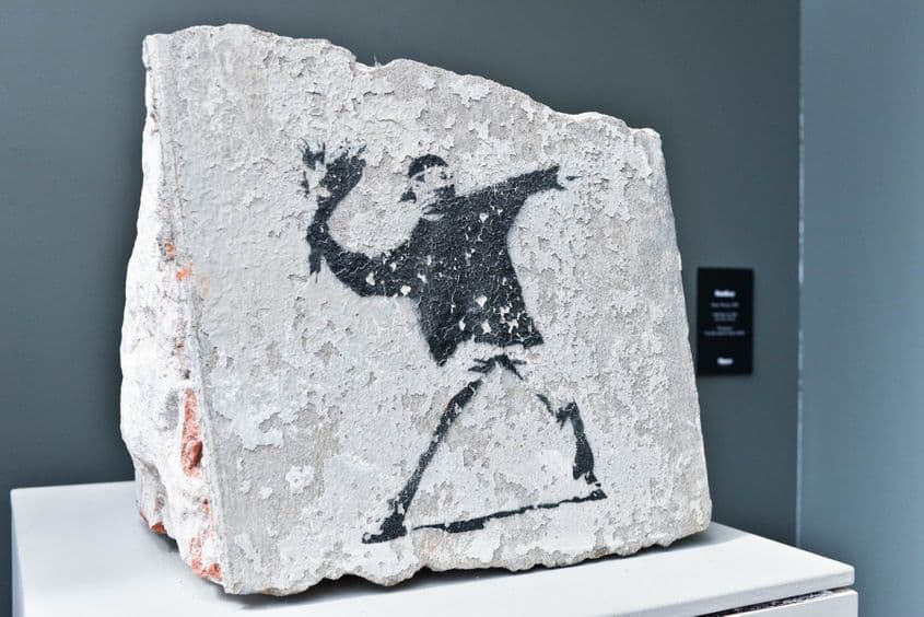 „Flower Thrower” (Aruncătorul de flori). Fragment de zid cu un graffiti creat de Banksy la Berlin în 2002, expus acum la Muzeul de artă MOCO din Amsterdam.